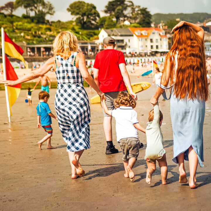 A family walks along a sandy beach in Lyme Regis, holding hands and enjoying a sunny day by the water.